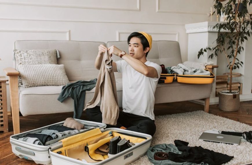 Man sitting on floor packing a suitcase, holding up a beige top, clothes and travel items spread around, beige sofa and plant in background, casual indoor setting