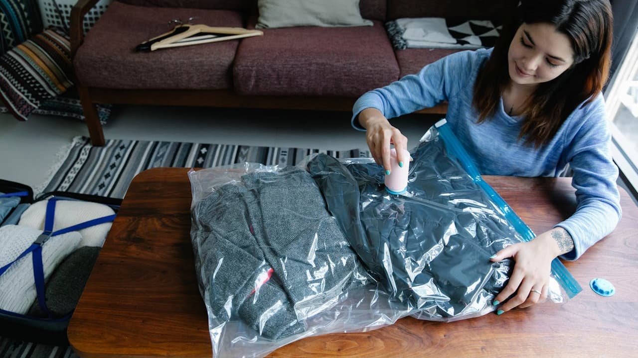 woman sealing clothes in vacuum storage bag, sitting at a wooden table, using a handheld vacuum, folded clothes compressed in plastic bag