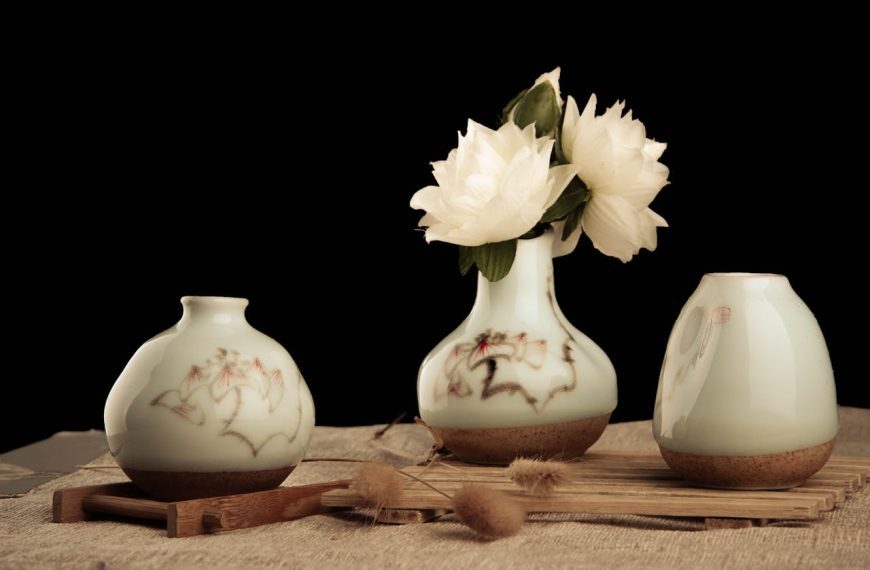 Three ceramic vases on a wooden surface, one vase holding white peony flowers, all vases decorated with subtle floral patterns, set against a black background