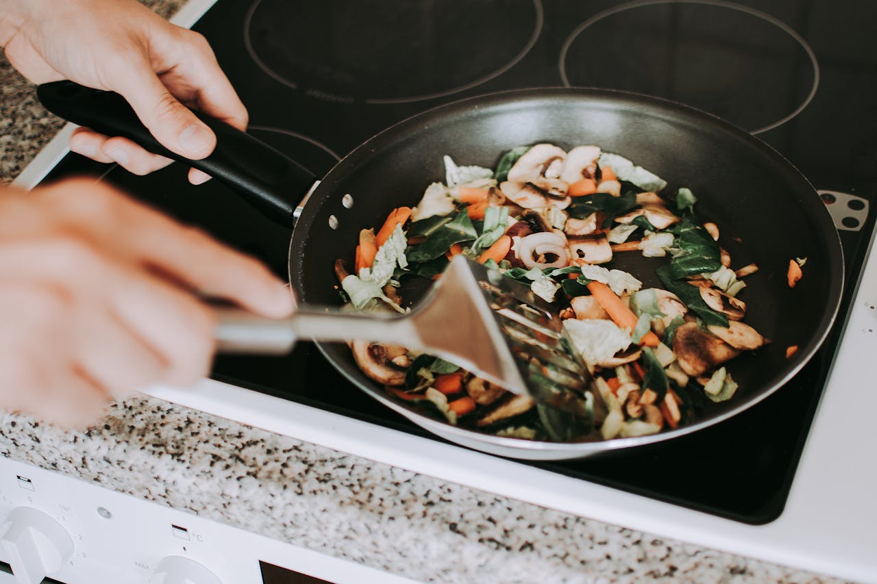 Close-up of a person stirring a colorful mix of vegetables, including mushrooms, carrots, and leafy greens, in a frying pan on a stovetop