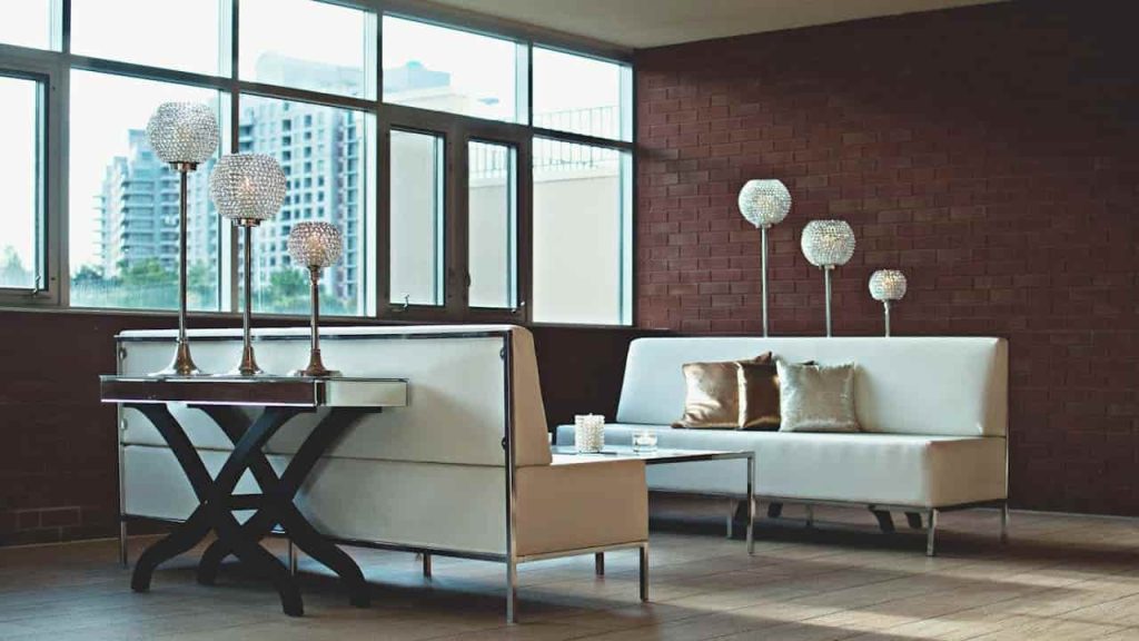 Modern lounge area with white leather sofas, velvet metallic-accent pillows, glass candle holders on a mirrored coffee table, and crystal orb lamps against a brick wall and large windows with city view