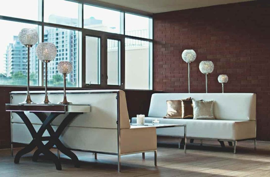 Modern lounge area with white leather sofas, velvet metallic-accent pillows, glass candle holders on a mirrored coffee table, and crystal orb lamps against a brick wall and large windows with city view