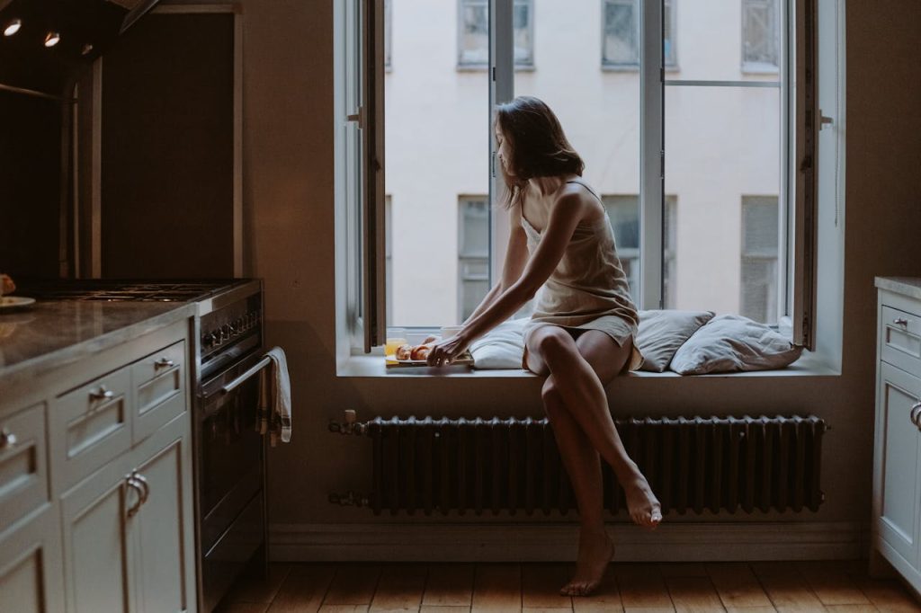 Woman sitting on a window seat in a dimly lit kitchen, reaching for a plate of croissants, with tall windows and industrial-style radiator below