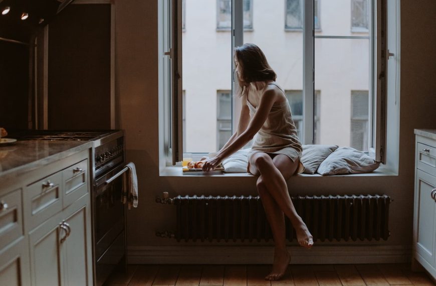 Woman sitting on a window seat in a dimly lit kitchen, reaching for a plate of croissants, with tall windows and industrial-style radiator below
