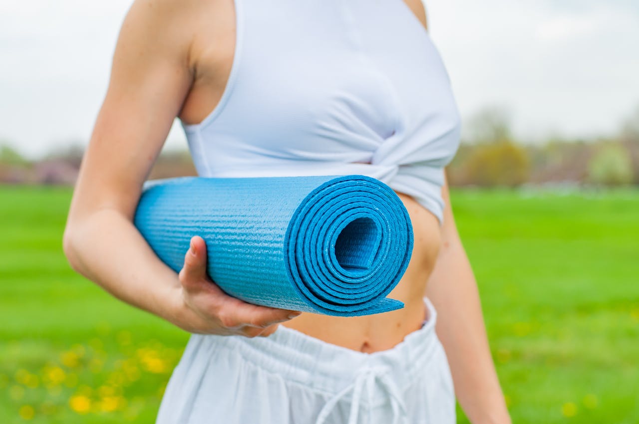 Person in white workout clothes holding a rolled blue yoga mat, standing outdoors on green grass, blurred park background