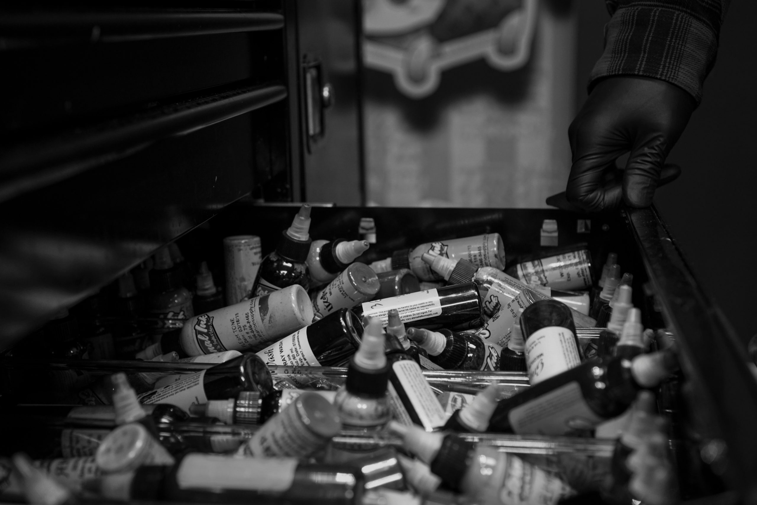 Black-and-white image of a gloved hand pulling open a drawer filled with assorted tattoo ink bottles, various labels and caps visible, stored in a metal cabinet