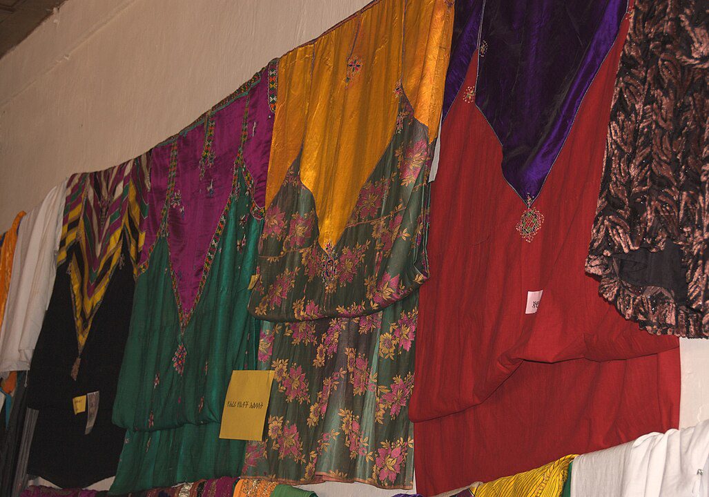 Colorful traditional garments hanging on display, including dresses in green, red, yellow, purple, and floral patterns, arranged side by side on a wall, showcasing intricate designs and fabrics