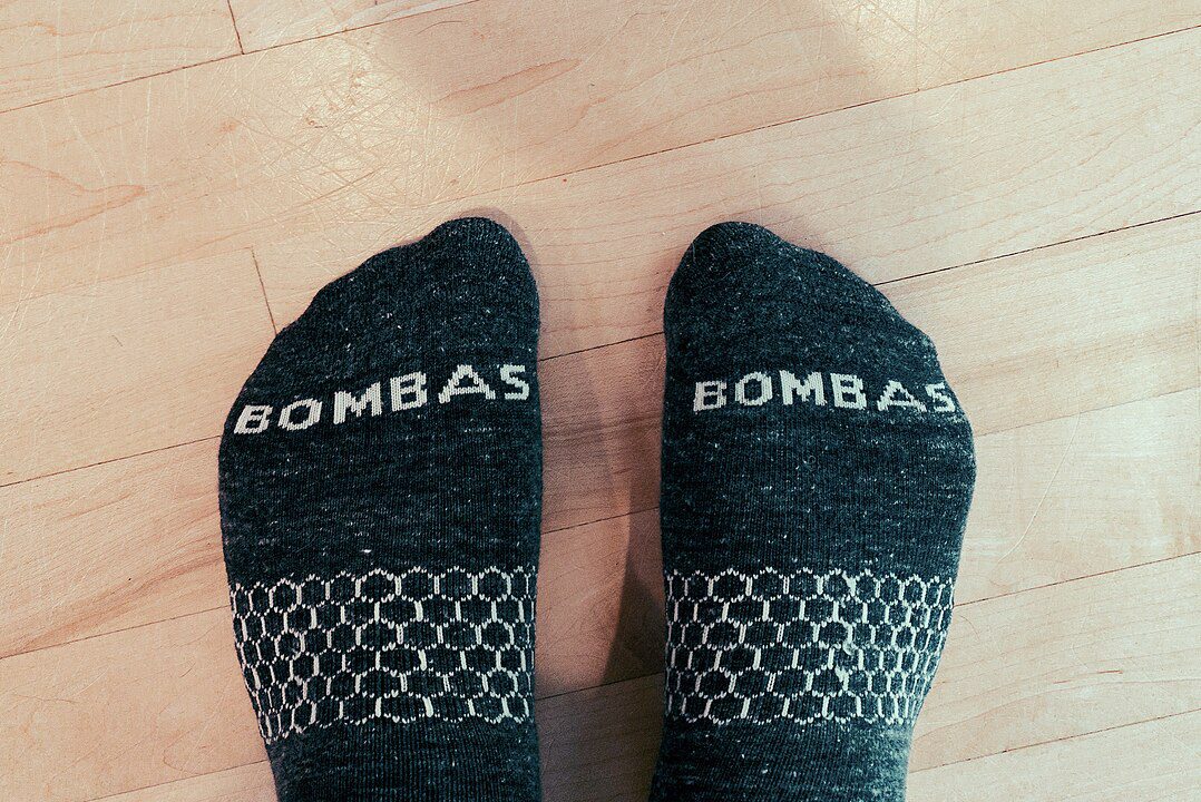 Close-up of two feet wearing black Bombas socks, white logo text on toes, geometric knit pattern on arch, standing on light wooden floor