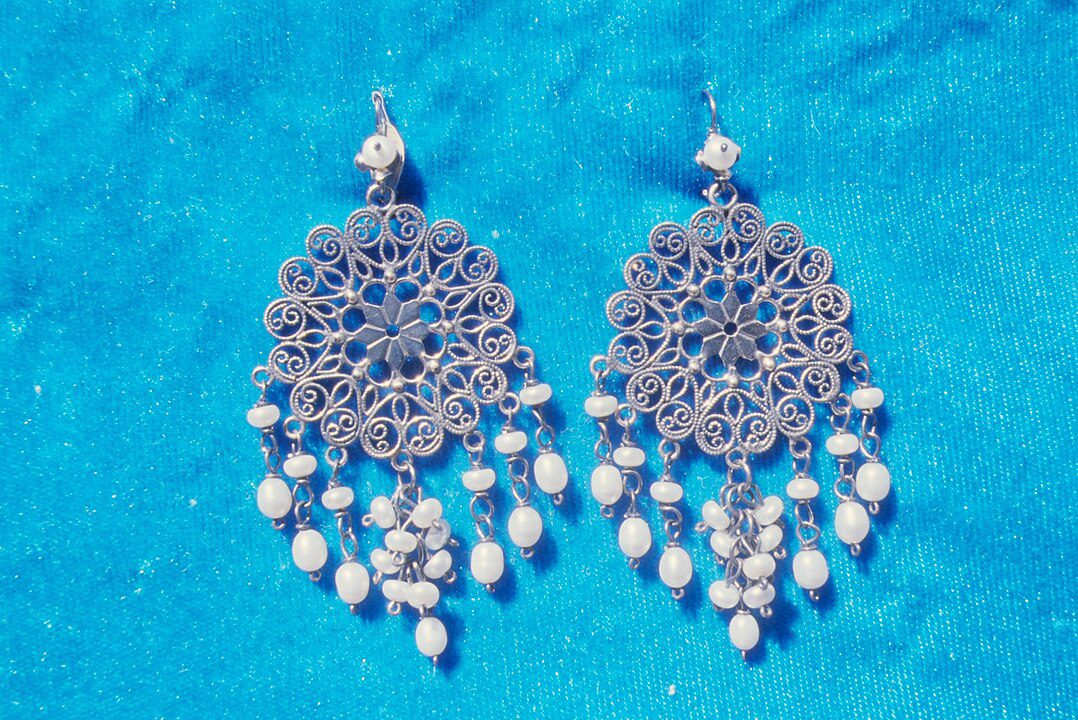 Pair of ornate silver chandelier earrings, detailed with intricate filigree work, adorned with small white pearls, displayed on a vibrant blue textured background