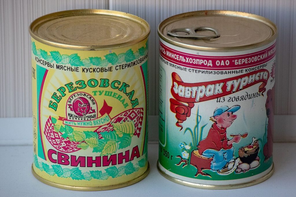 Two vintage canned meat tins with colorful illustrations, the left labeled "Astrocolora Cinnamon" with a floral and character design, the right labeled "Rindfleisch Frühstücksfleisch" featuring a boy and bear cartoon, both with gold lids, placed side by side on a light surface