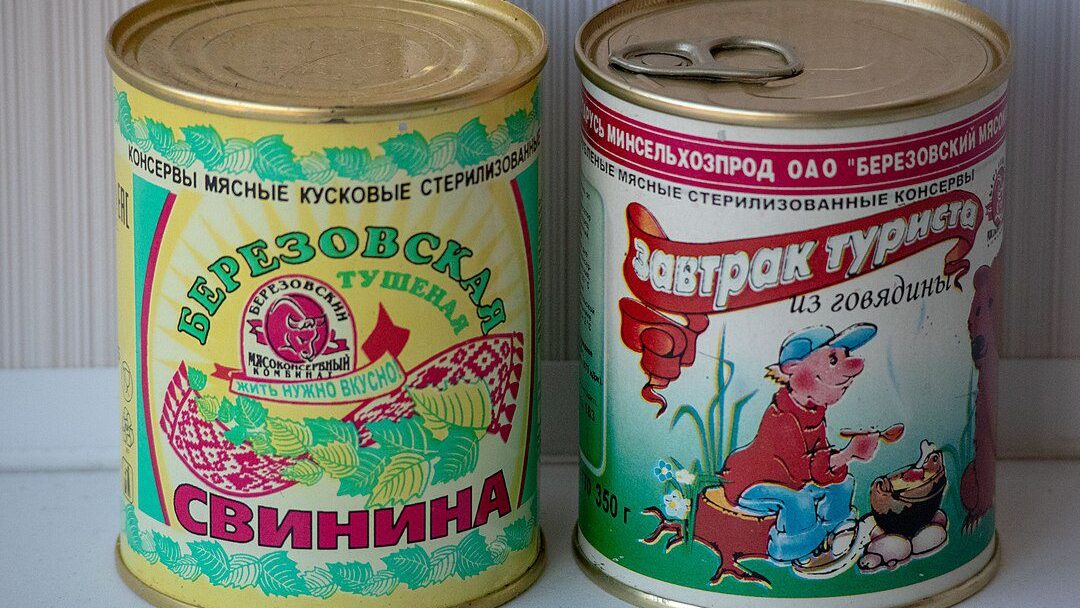 Two vintage canned meat tins with colorful illustrations, the left labeled "Astrocolora Cinnamon" with a floral and character design, the right labeled "Rindfleisch Frühstücksfleisch" featuring a boy and bear cartoon, both with gold lids, placed side by side on a light surface