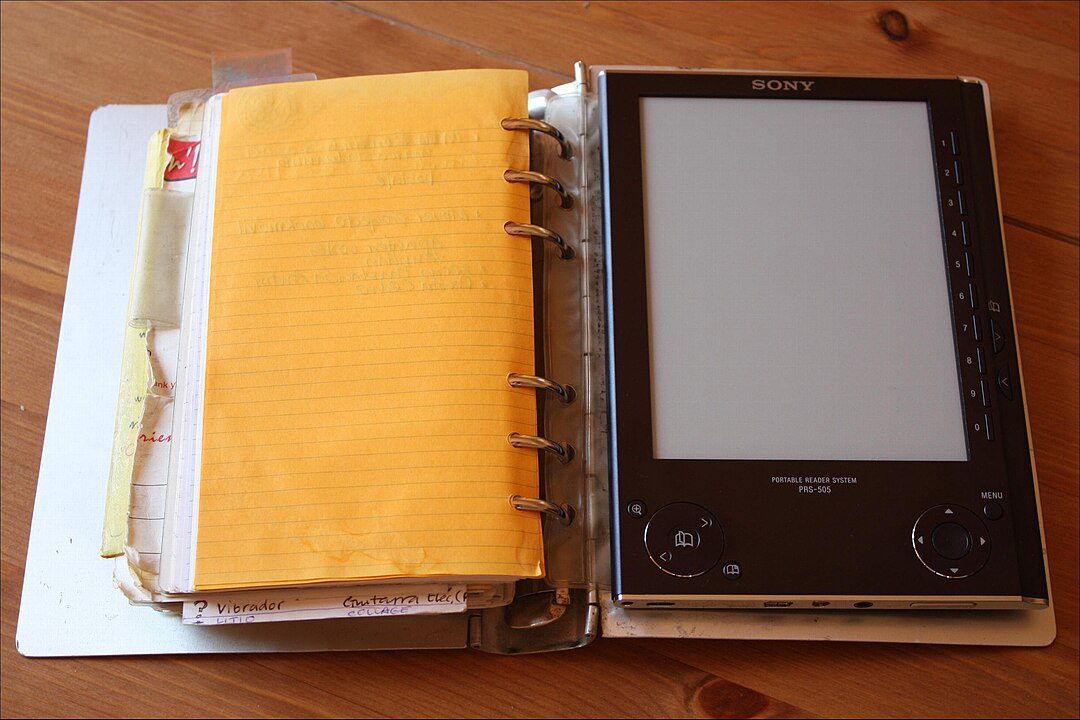 Open binder with yellow paper on the left, e-reader or tablet device on the right, placed on a wooden surface