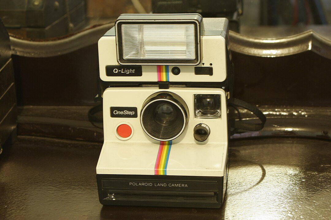 Vintage Polaroid instant camera, white body with rainbow stripe, large flash unit on top, black lens in center, red shutter button, placed on a polished wooden surface
