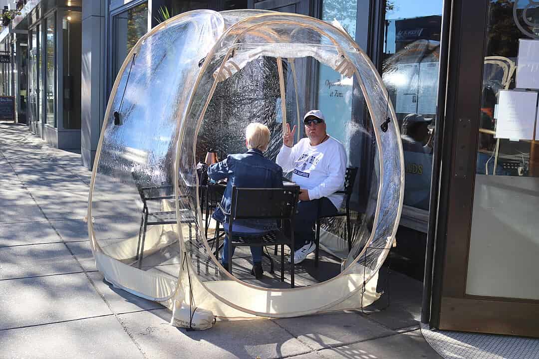 Plastic dining bubble, transparent dome structure, enclosed around restaurant table and chairs, zippered entrance, outdoor patio setting, weather-resistant material, spaced apart for social distancing