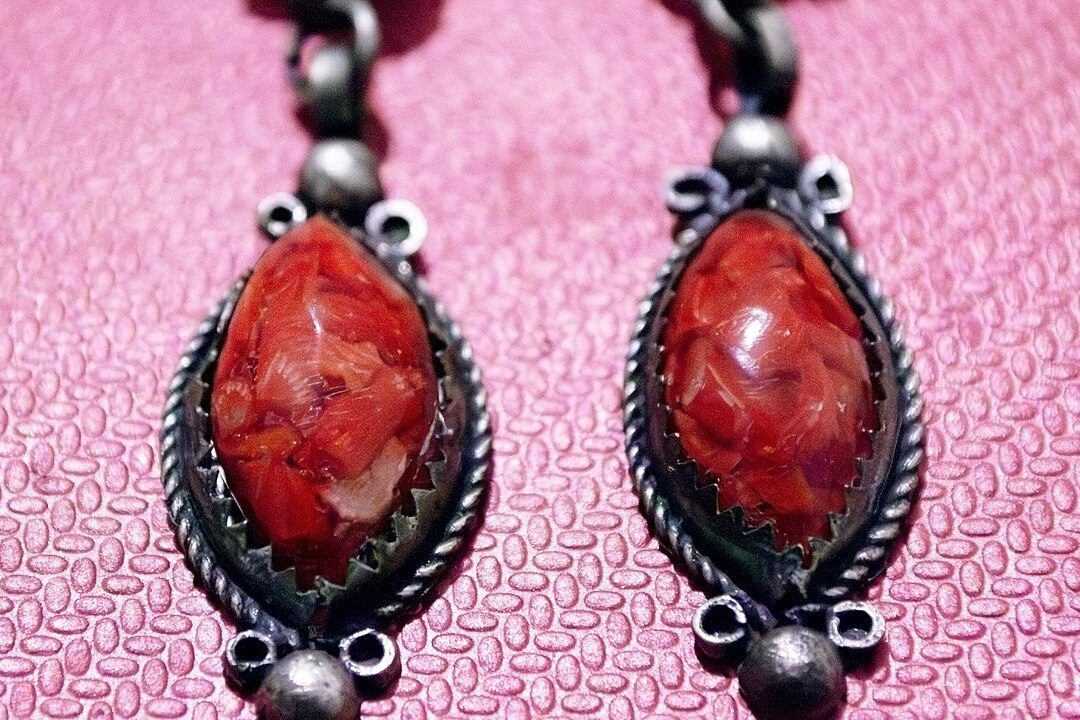 Close-up of two vintage-style earrings with oval red-orange stones, set in detailed silver-toned metal frames, small metallic beads at the top and bottom of each earring, displayed on a pink textured fabric background
