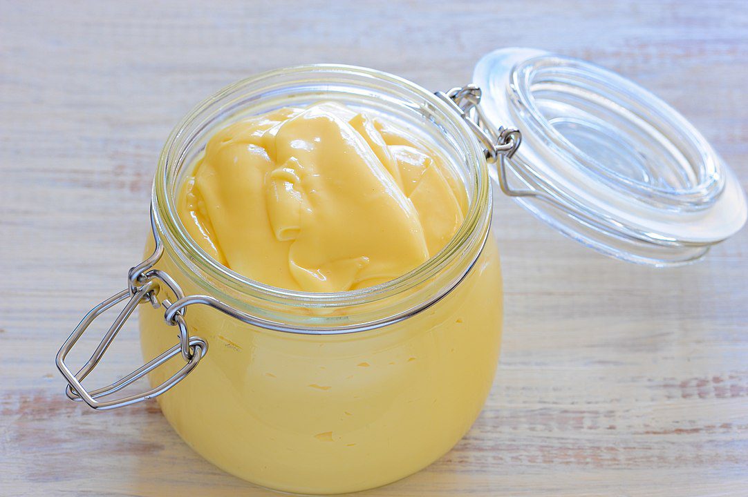 Glass jar filled with smooth, creamy yellow custard or lemon curd, placed on a light wooden surface, with the hinged lid open