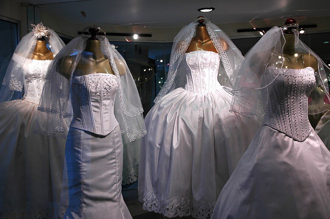 Display of five mannequins wearing white wedding dresses, each with different styles and veils, arranged in a store window, illuminated under soft lighting