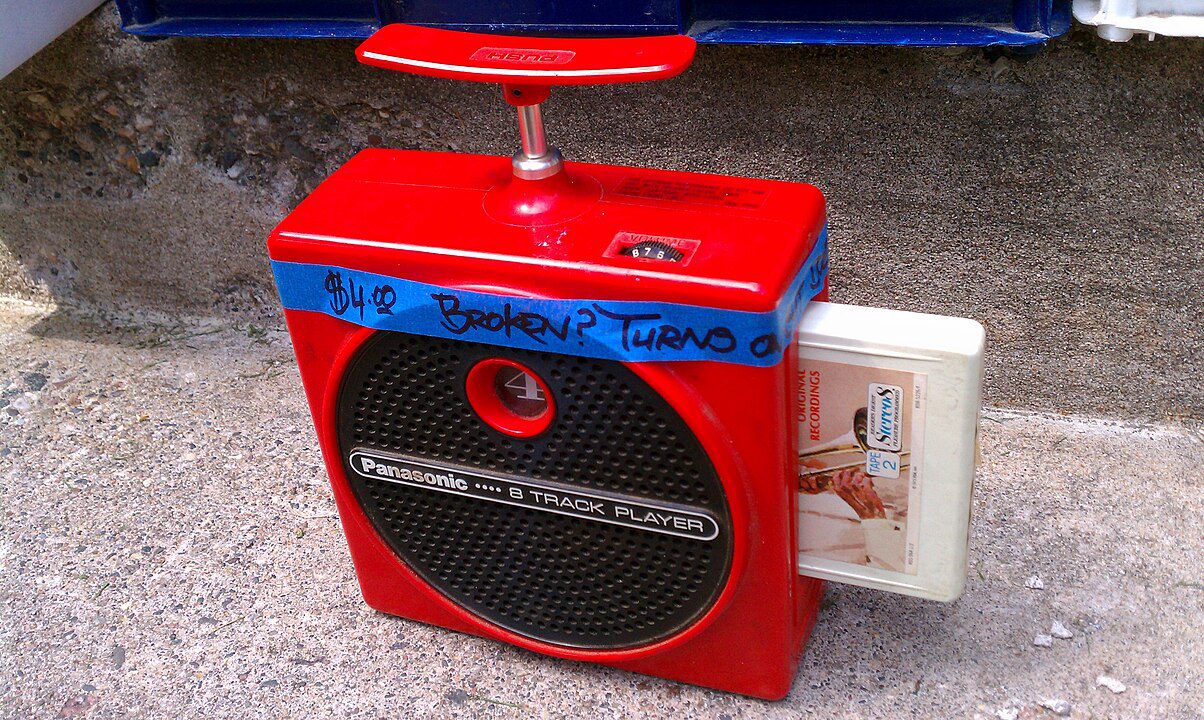 Red vintage 8-track player with a black circular speaker area, white 8-track cartridge inserted on the side, blue tape across the top, placed on a concrete surface