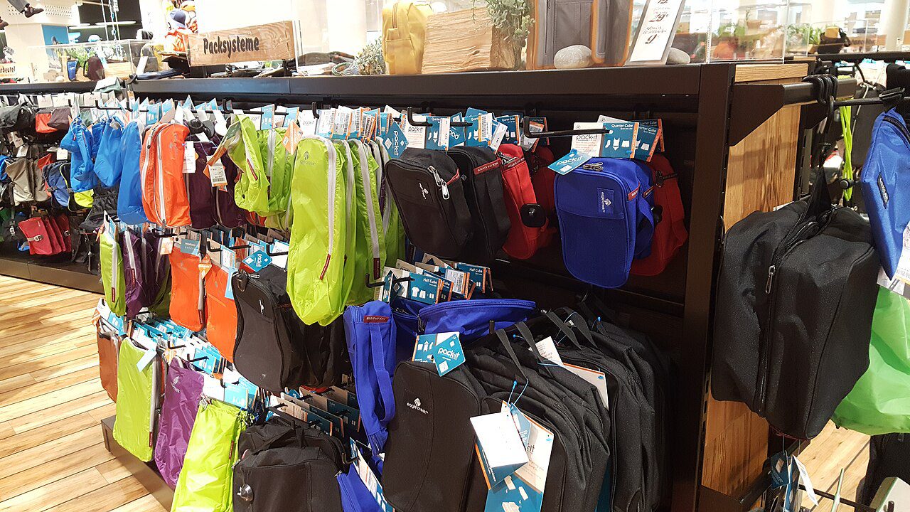 Multiple colorful packing cubes, hanging on display racks, arranged by size and color, indoor retail store setting, product tags attached, wooden flooring beneath, travel accessories section