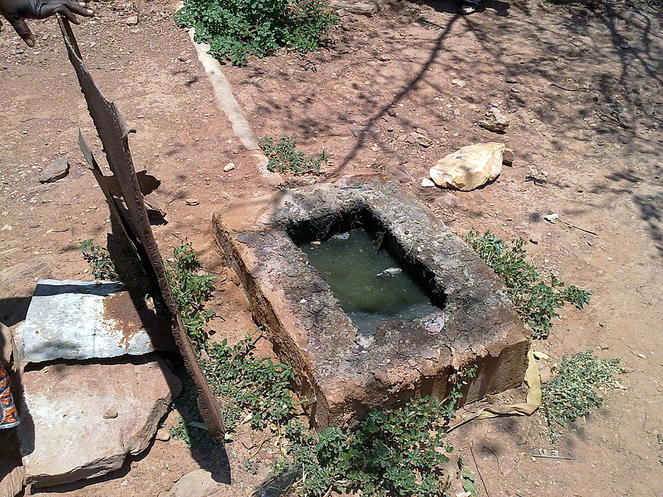 damaged septic tank, open concrete structure, standing dirty water, located in sandy soil, surrounded by dry vegetation and debris