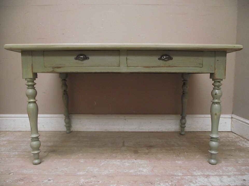 Old wooden table, green paint finish, two drawers with round knobs, turned legs, placed against a beige wall, sitting on a worn carpet