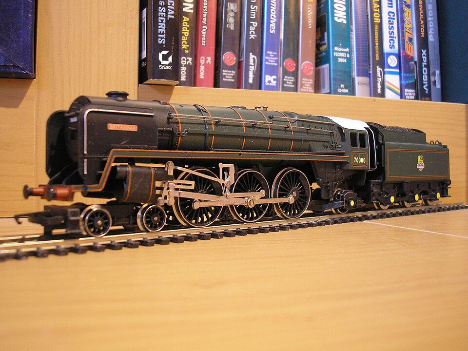 Model steam locomotive train, black with detailed parts, displayed on a wooden shelf, background of colorful upright books, miniature railway setting, realistic design and finish