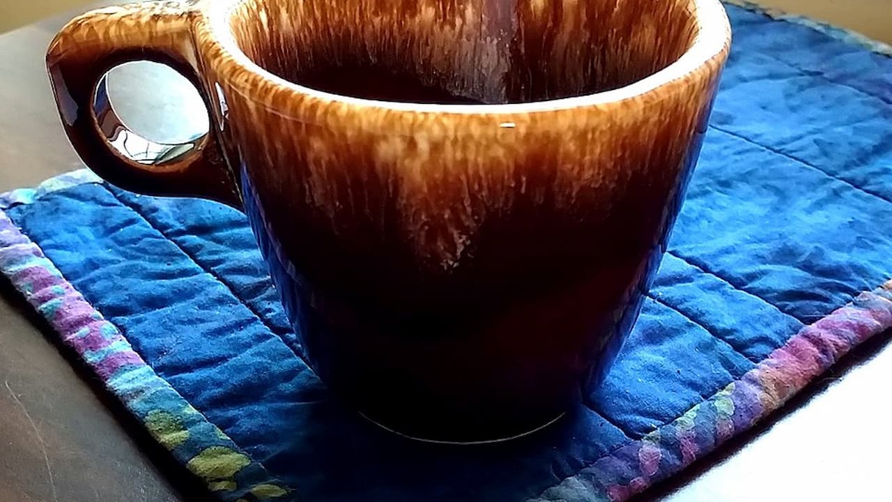 Brown ceramic mug with a glossy, drip-like glaze near the rim, sitting on a textured blue quilted coaster with colorful trim, placed on a wooden surface