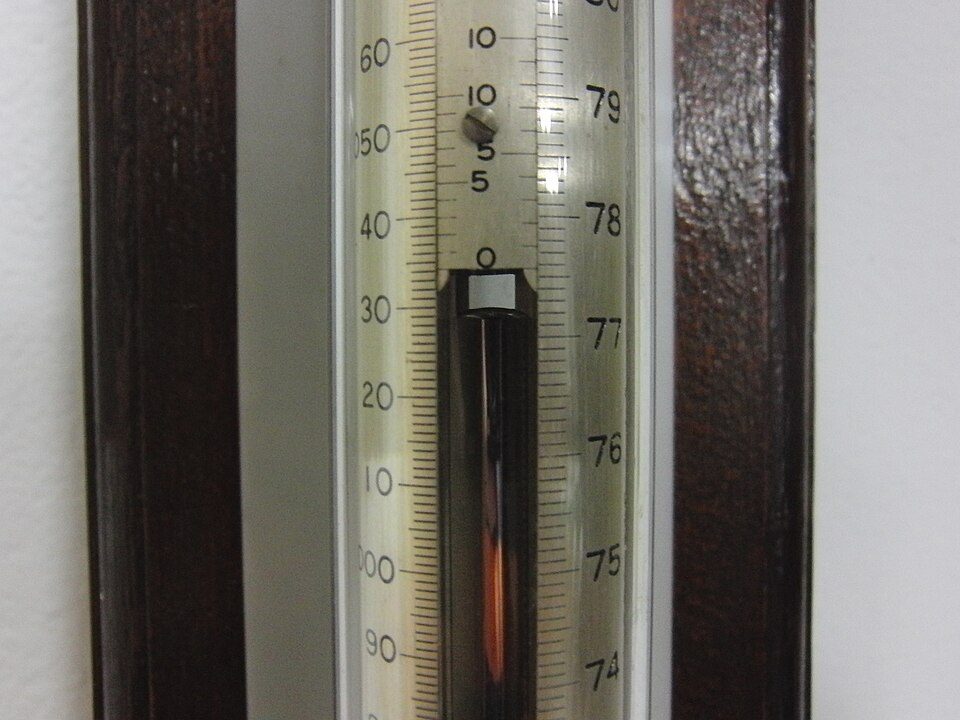 Close-up of a wall-mounted thermometer, white background with black markings, displaying temperature around 26°C, mercury visible inside glass tube, wooden surface in the background