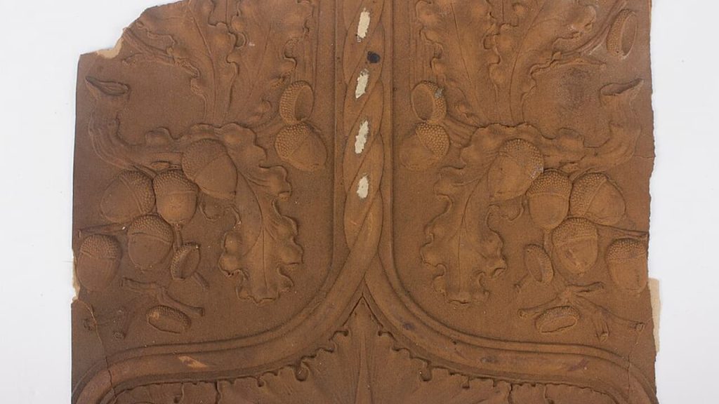 Aged wallpaper scrap, brown with intricate symmetrical floral and vine patterns, slightly torn edges, visible vertical seam, vintage or antique design