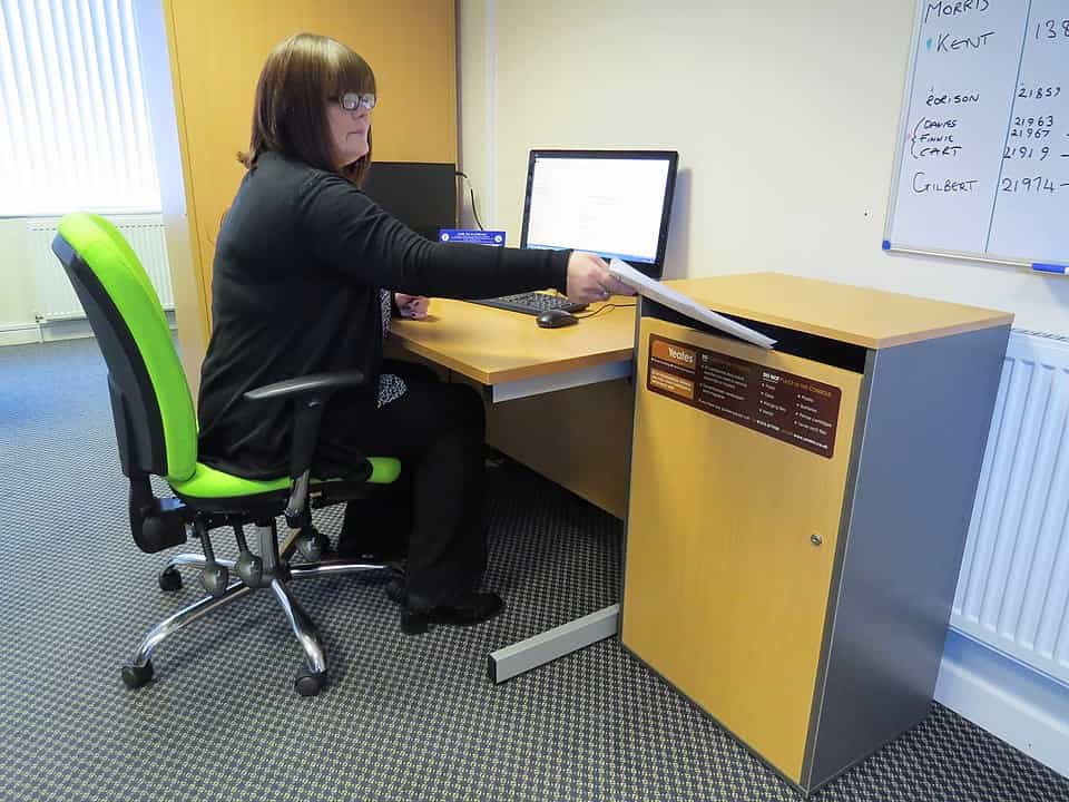 Woman shredding old documents, standing beside electric shredder, holding paper over machine slot, office setting, waste bin partially filled with shredded paper, casual clothing, focused expression