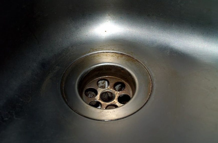 soap scum in kitchen sink drain, white residue buildup, greasy film, cloudy drain edges, metal surface stained, partially clogged appearance