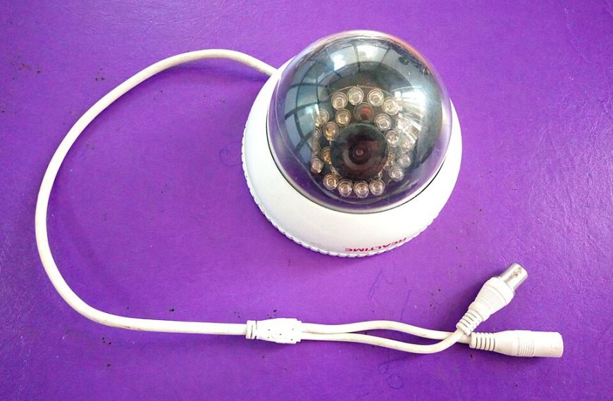 White dome CCTV security camera, visible lens and infrared LEDs, attached white power and video cables, placed on a solid purple surface