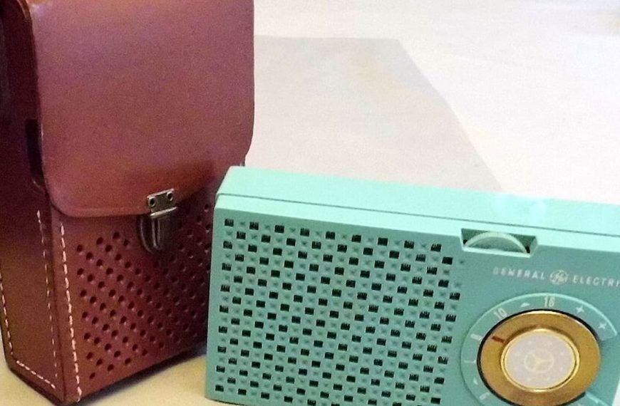 Vintage turquoise transistor radio, circular speaker grill on the front, tuning dial on top, paired with a brown leather carrying case, both placed on a light-colored surface, retro design and compact size