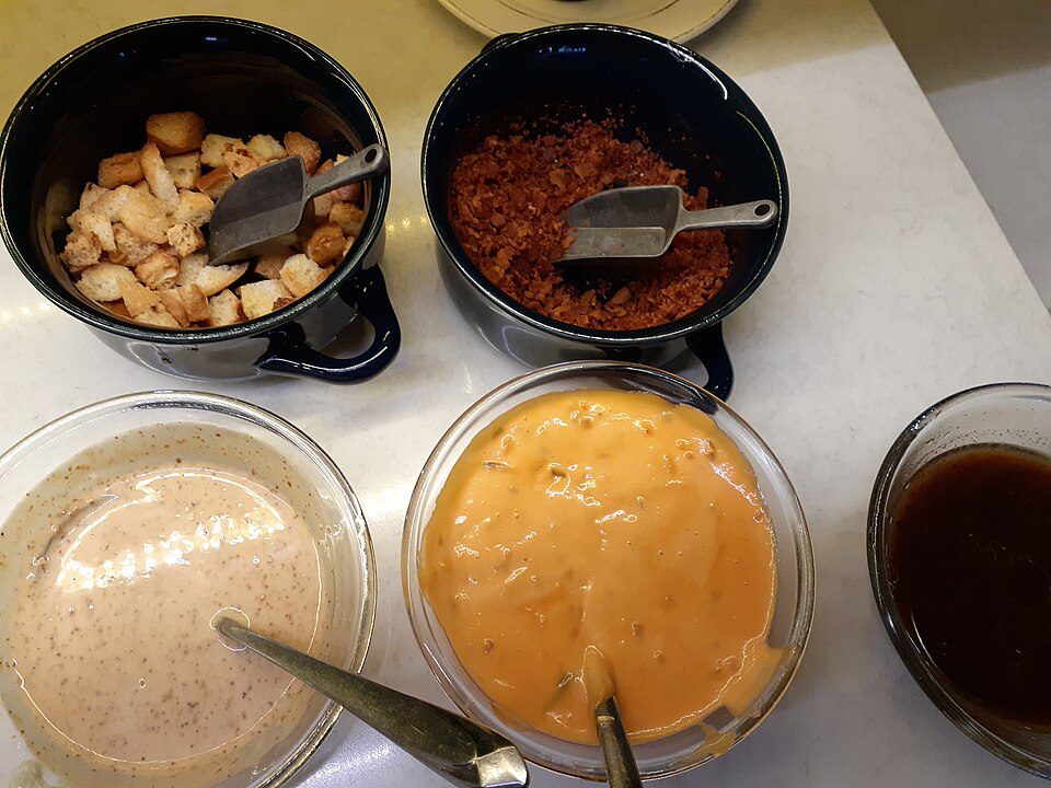 assortment of pizza toppings and sauces, diced chicken in a black bowl, crumbled bacon in a black bowl, creamy white sauce in a clear bowl, orange cheese sauce in a clear bowl, dark liquid in a glass, spoons and knives for serving, ingredients arranged on a white surface