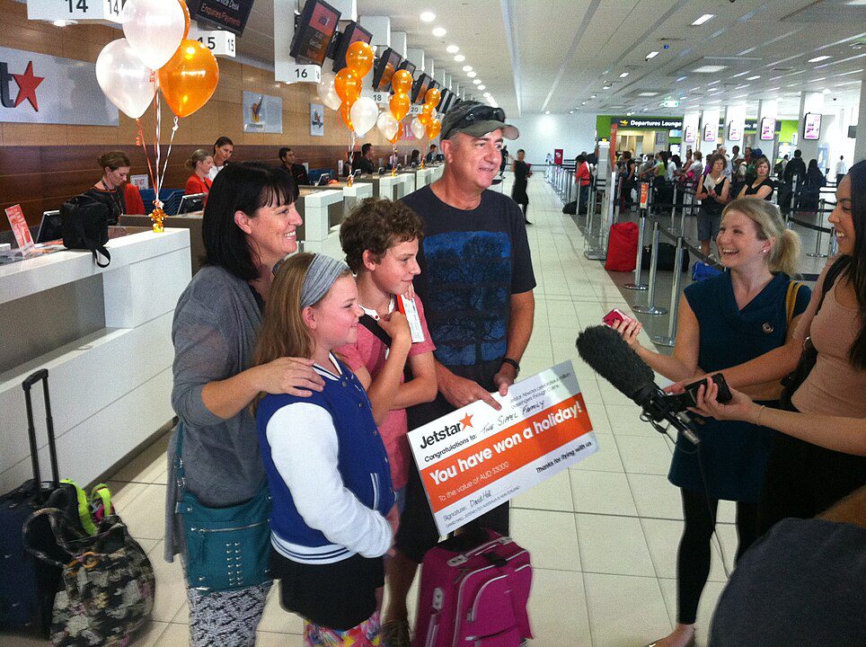 Group of people at an airport check-in area, holding a large orange and white travel voucher, smiling, surrounded by luggage and airline staff, media personnel recording the moment