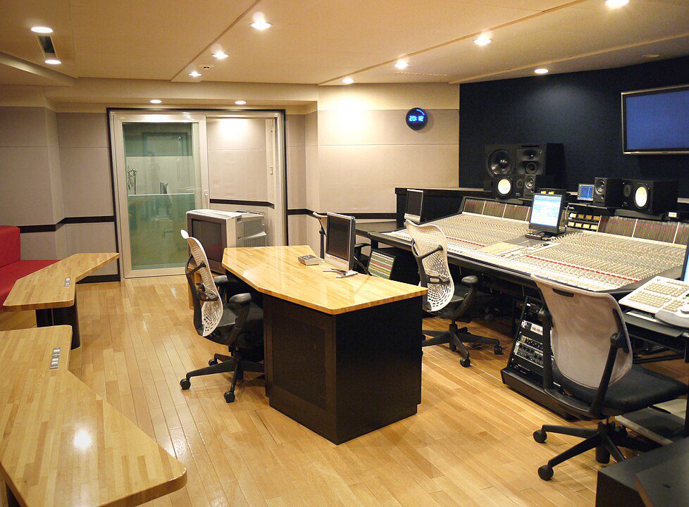 modern recording studio, multiple office chairs, long wooden desks with audio equipment and monitors, soundproof walls, wooden floor, overhead lighting, glass door in the back