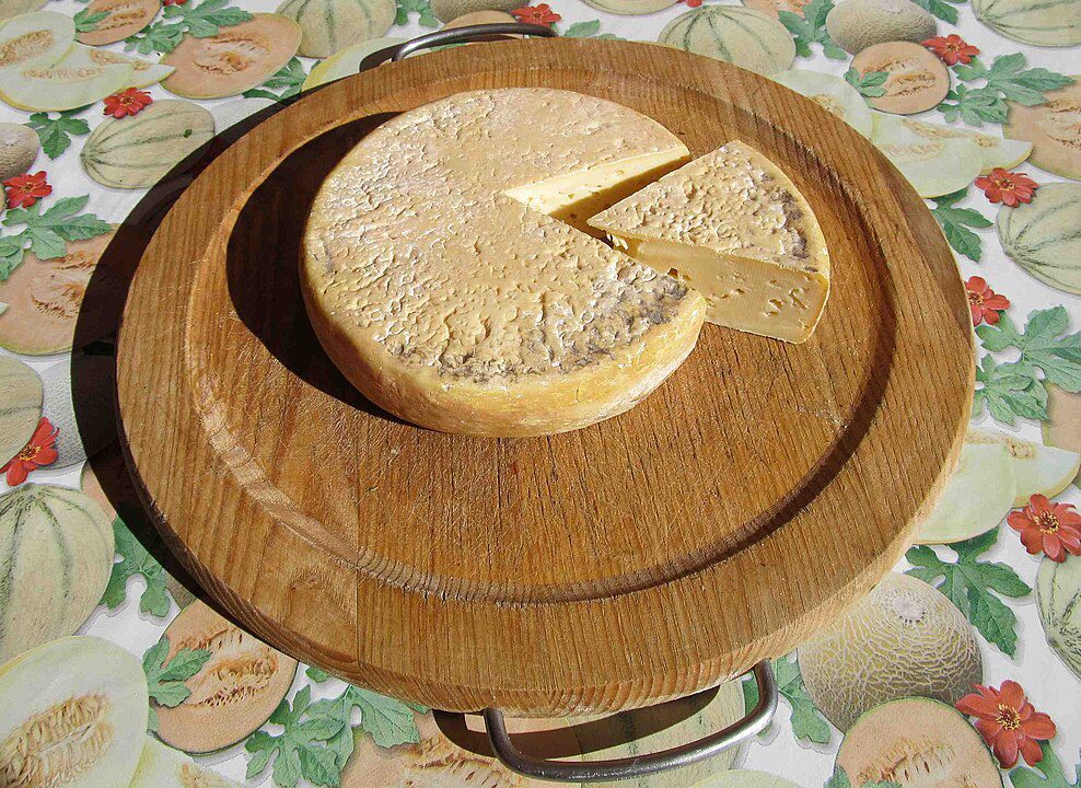 Round cheese wheel on a wooden plate, one wedge sliced, placed on a festive tablecloth, textured cheese surface, rustic food presentation