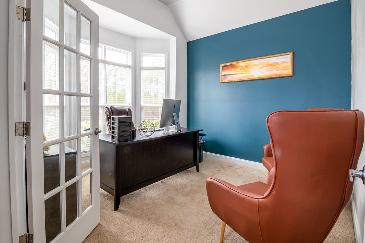 Modern home office with a bold teal accent wall, large windows, and minimalist dark wood furniture