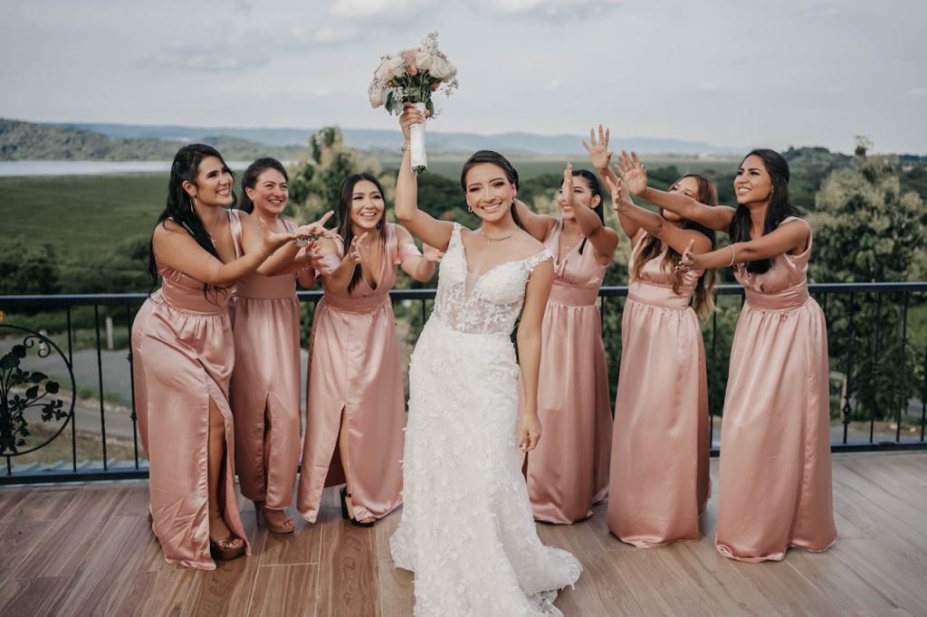 Bride in white wedding gown holding bouquet up as bridesmaids in pink dresses reach toward her, smiling outdoors