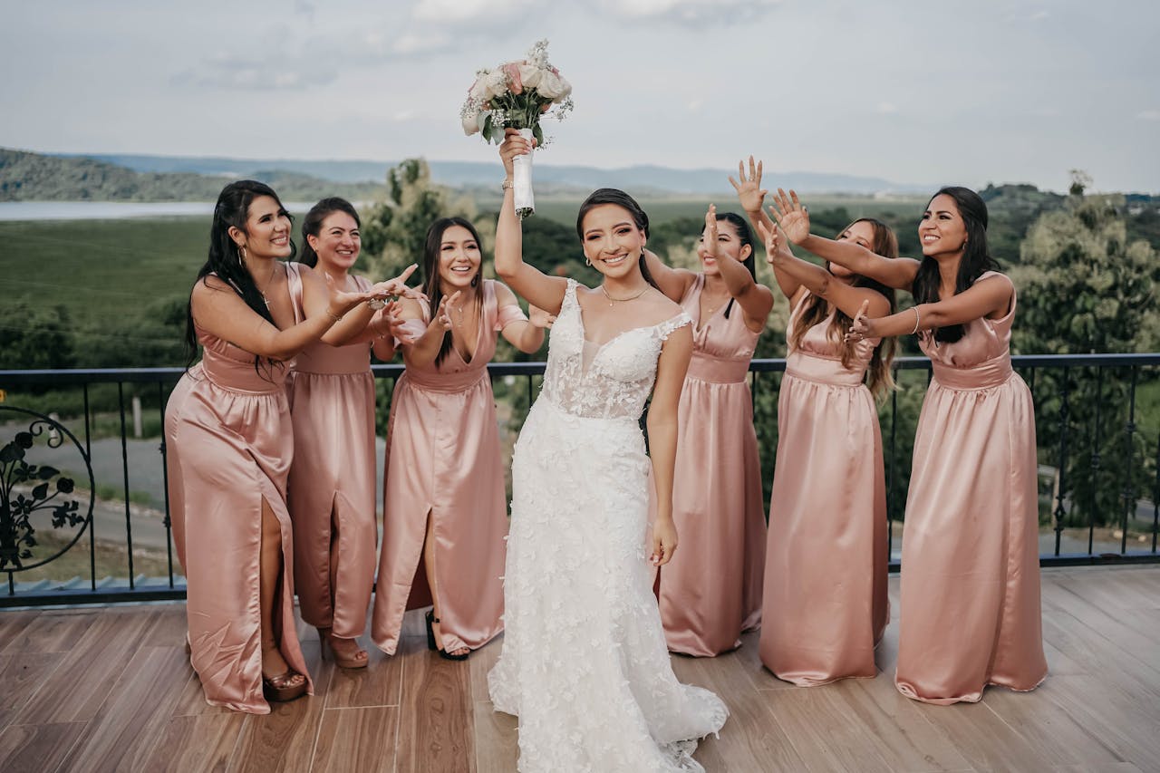 Bride in white wedding gown holding bouquet up as bridesmaids in pink dresses reach toward her, smiling outdoors