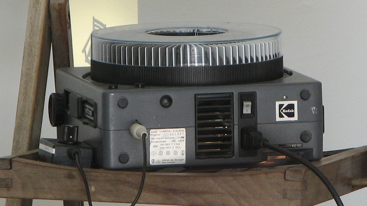 Vintage slide projector with a circular slide tray on top, placed on a wooden surface, various input and control ports visible on the back, black power cable plugged in, neutral background