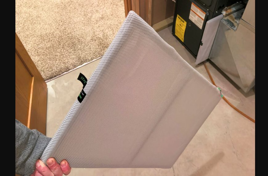 Person holding clean HVAC air filter with white mesh cover, standing near furnace in residential utility room