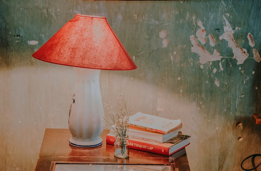White plastic table lamp with red shade on wooden nightstand, beside stacked books and a small vase.