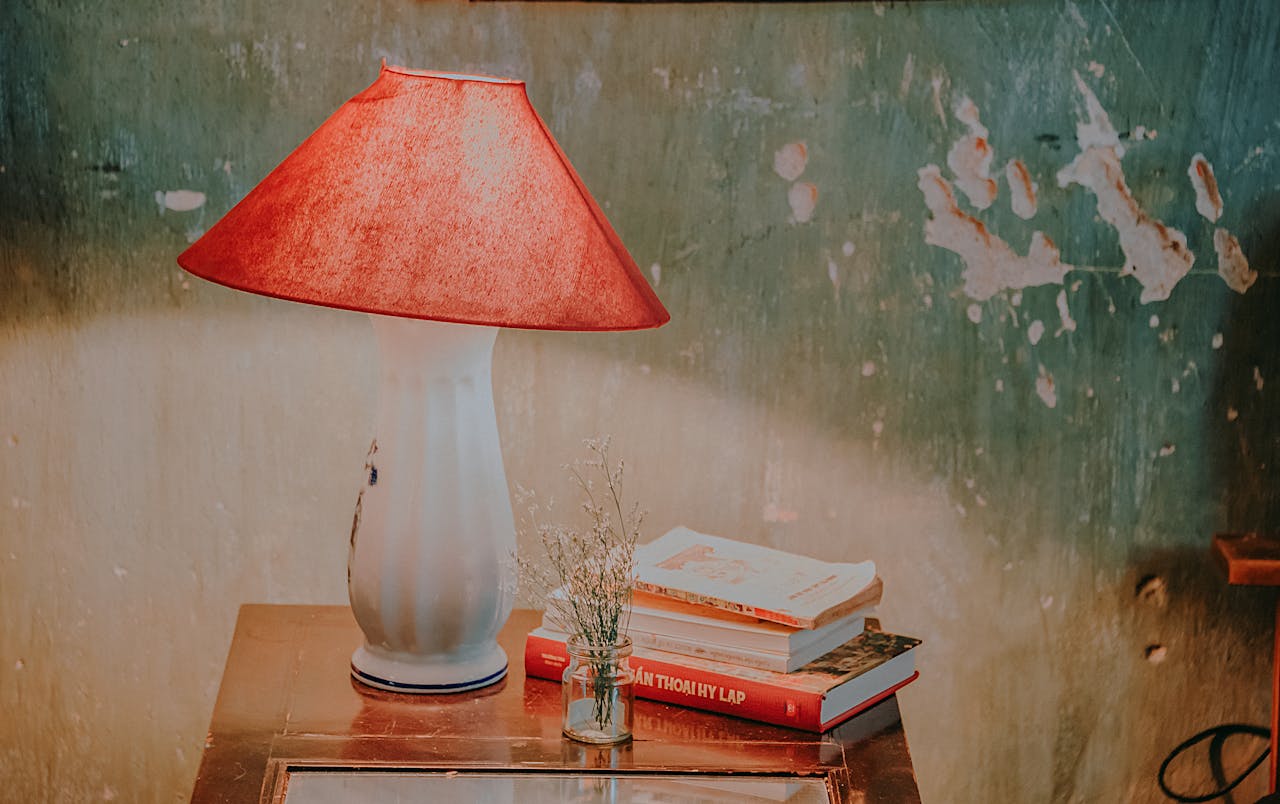 White plastic table lamp with red shade on wooden nightstand, beside stacked books and a small vase.