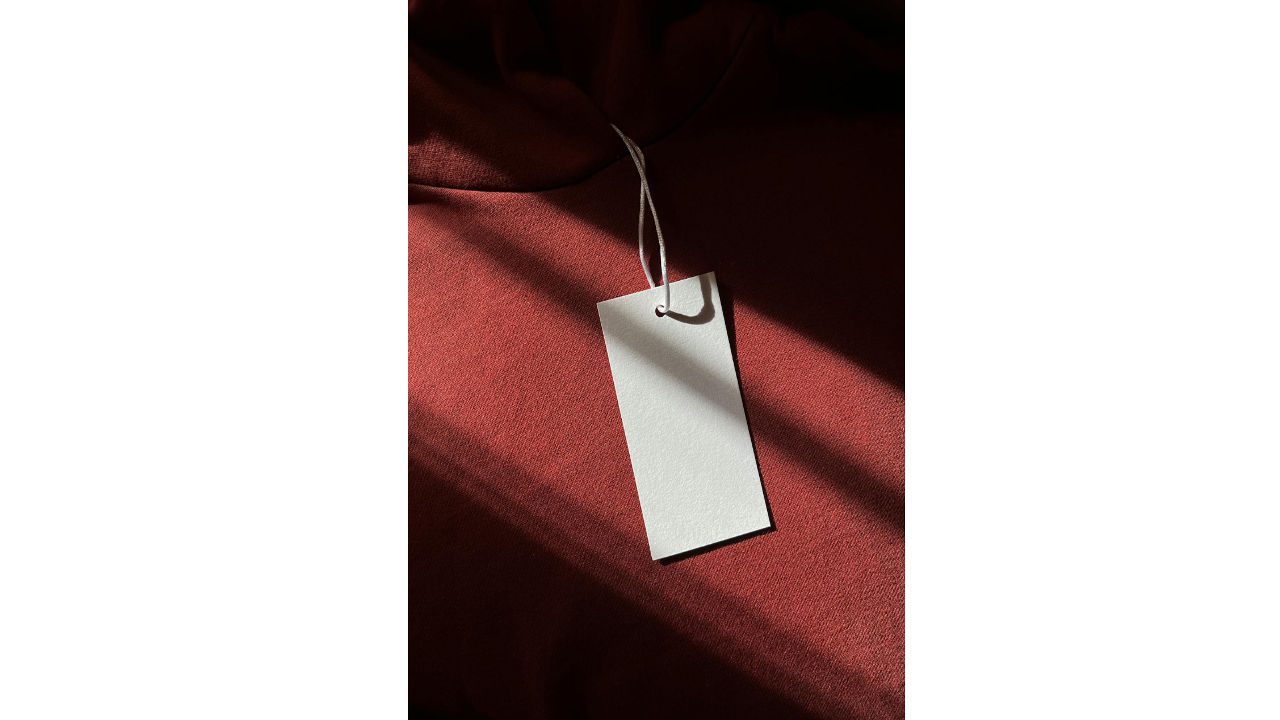 White blank clothing tag with string resting on maroon fabric in sunlight