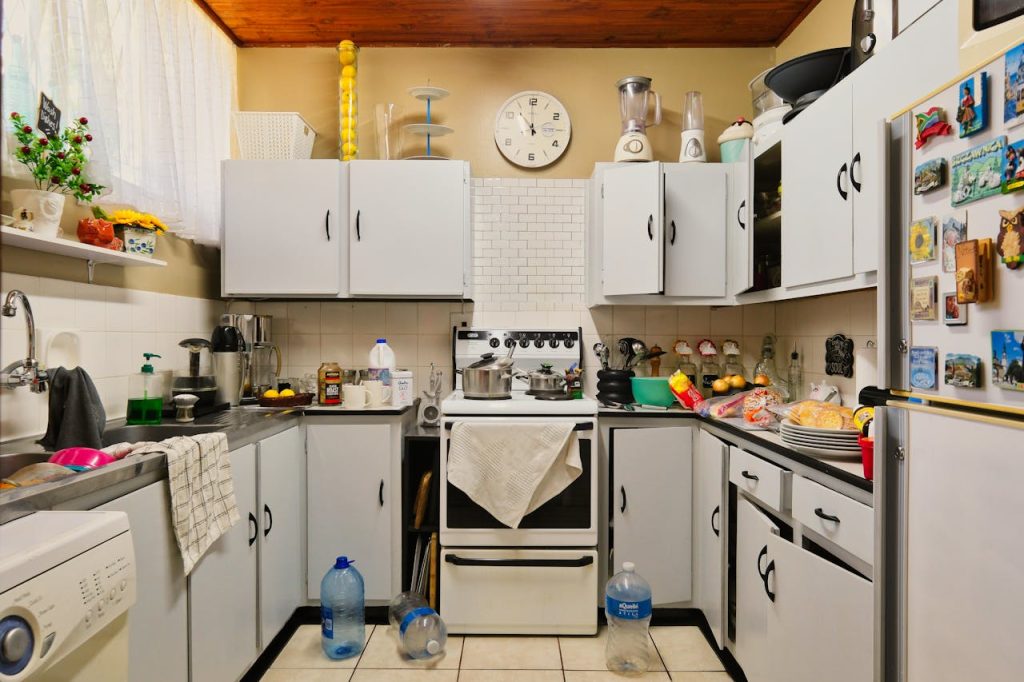 Cluttered kitchen with white cabinets, dishes and utensils on countertops, pots on stove, scattered bottles on floor, refrigerator with colorful magnets