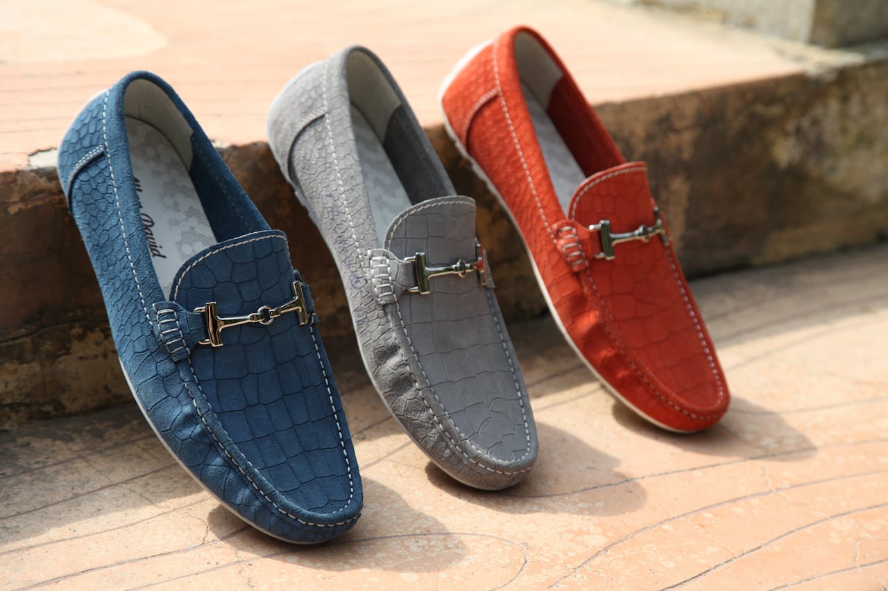 hree pairs of crocodile-textured loafers in blue, gray, and red displayed outdoors