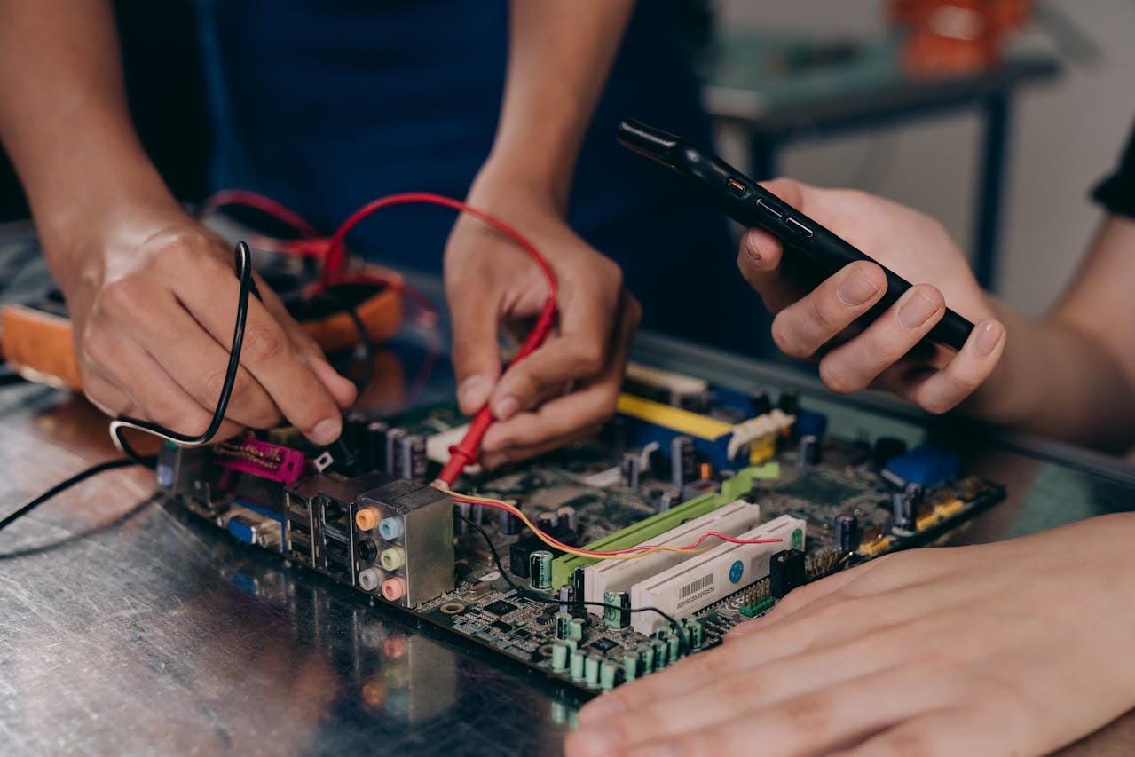 Hands working on computer motherboard with multimeter probes, person holding smartphone, electronic repair setup