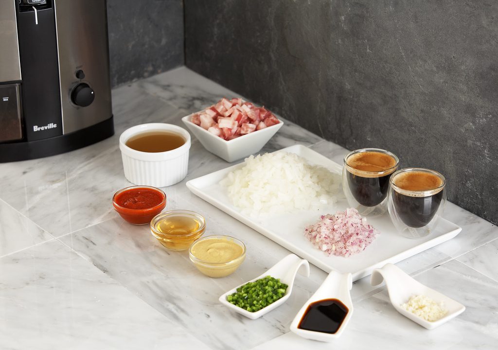 Ingredients arranged on a kitchen counter, including a bowl of chopped vegetables, a plate of cooked white rice, chopped onions, chopped garlic and ginger, red chili sauce, green sauce, soy sauce, two cups of dark liquid (possibly coffee or broth), and a bowl of light brown sauce or stock, with a cooking appliance in the background