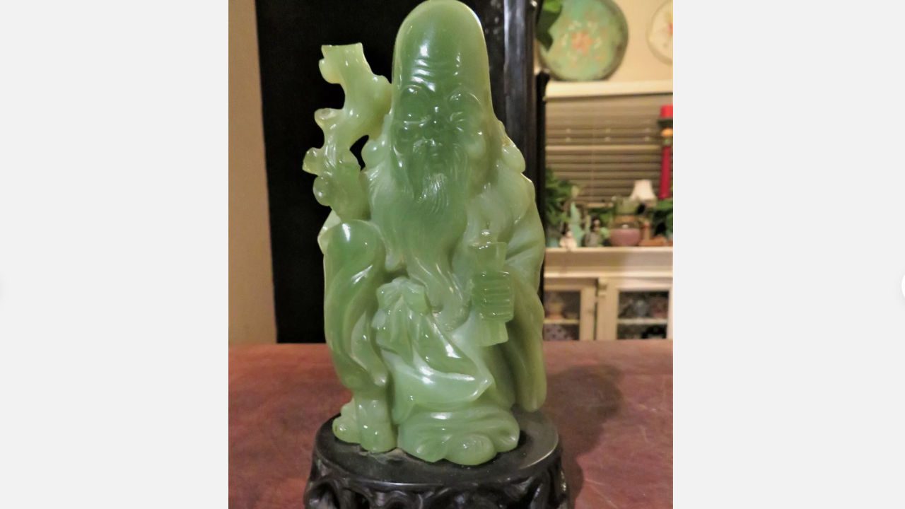 green jade carving of elderly man with long beard, holding staff and scroll, displayed on black ornate base, background shows indoor shelf with decorative items
